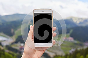 Outdoor image of one hand holding and showing white smartphone with blank black desktop screen with blur green mountains on