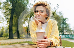 Outdoor image of pretty woman drinking coffee at terrace cafetaria in the park. Female in the city street drinking hot cappuccino