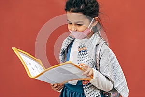 Outdoor image of smiling child wearing protective face mask looking in a folder with papers after school. Pupil little girl in