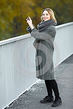 Outdoor lifestyle photo of young beautiful blonde woman in fall autumn park taking photos on smartphone cozy scarf grey vintage co