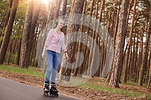 Outdoor photo of attractive magnetic woman having positive facial expression, having roller skates on legs, listening to music,