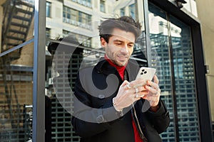 Outdoor photo of young brown haired unshaved guy keeping mobile phone in raised hands and looking positively on screen while