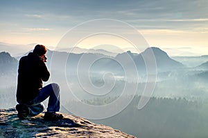 Outdoor photographer with tripod and camera on rock thinking. Thick fog in autumnal countryside. Man work on sharp clif