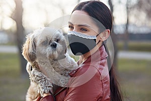 Outdoor picture of delighted lovely young female wearing antibacterial mask to protect from coronavirus, holding white Maltese dog