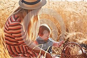 Outdoor picture of young fair haired mother sitting with her little daughter at wheat field, having small picnic together, mum