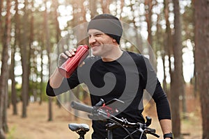 Outdoor picture of young man wearing dark tracksuit, having happy facial expression, laughing sincerely, holding red bottle with