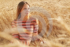 Outdoor portrait of adorable beautiful woman sitting on ground at wheat field, looking aside, having pleasant facial expression,