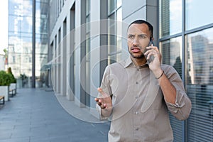 Outdoor portrait of angry and upset man outside office building, businessman in shirt talking on phone and gesturing