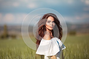 Outdoor portrait of attractive young woman with long wavy hair wear white dress over green grass field at sunset