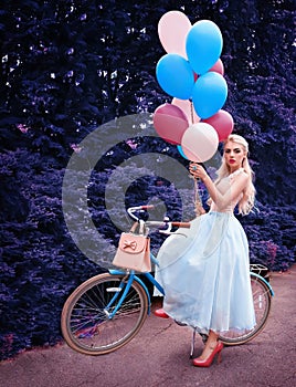 Outdoor portrait of a beautiful blonde girl holding balloons and riding a bike
