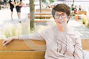 Outdoor portrait of beautiful and elegant middle age 55 - 60 year old woman wearing long sleeve top sitting on bench on