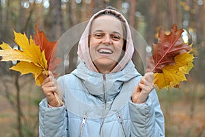 Outdoor portrait of cute extremely happy smiley woman holding autumn leaves in the nature, looking at camera and laughing enjoying