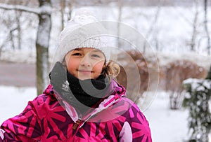 Outdoor portrait of cute little girl in white knitted hat in winter park smiling looking at camera.