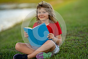 Outdoor portrait of a cute little kid reading a book in summer park. Blond kid boy sitting on grass and reading book