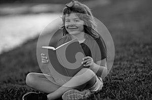 Outdoor portrait of a cute little kid reading a book in summer park. Blond kid boy sitting on grass and reading book