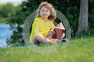 Outdoor portrait of a cute little kid reading a book in summer park. Blond kid boy sitting on grass and reading book