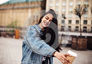 Outdoor portrait of a dark-haired young woman in a denim jacket holds coffee to go. Emotional woman posing with happy smile on the