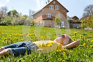 Outdoor portrait of European boy lies on grass in dandelions flower filed looking to the sky on against house. Side view