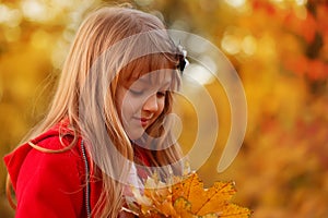 Outdoor portrait of happy blonde child girl in a red jacket holding yellow leaves. Little girl walking in the autumn park or