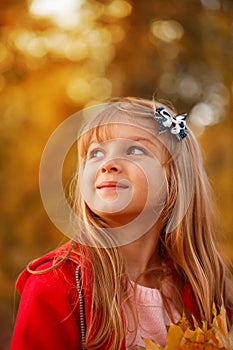 Outdoor portrait of happy blonde child girl in a red jacket holding yellow leaves. Little girl walking in the autumn park or