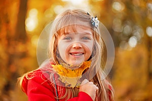 Outdoor portrait of happy blonde child girl in a red jacket holding yellow leaves. Little girl walking in the autumn park or