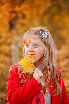 Outdoor portrait of happy blonde child girl in a red jacket holding yellow leaves. Little girl walking in the autumn park or