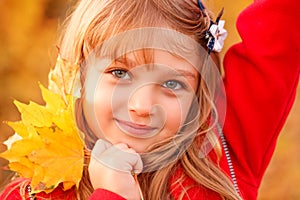 Outdoor portrait of happy blonde child girl in a red jacket holding yellow leaves. Little girl walking in the autumn park or