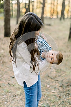 Outdoor portrait of happy pretty mother holding on hands her cute little son, enjoying their joint walk in pine forest