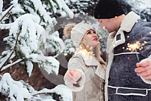 Outdoor portrait of happy romantic couple celebrating Christmas with burning fireworks in snowy forest.