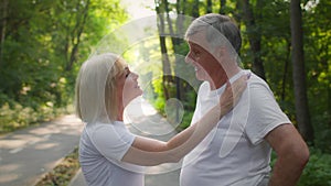 Outdoor portrait of happy senior couple in love talking during walk in green public park, woman caressing her husband