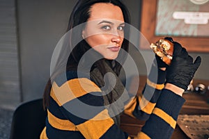 Outdoor portrait of hungry pretty woman wearing casual sweater, eatting delicious burger while sitting at terrace. Female with red