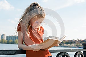 Outdoor portrait of little schoolgirl with book, girl child 7, 8 years old with glasses reading textbook