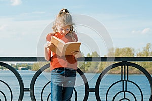 Outdoor portrait of little schoolgirl with book, girl child 7, 8 years old with glasses reading textbook