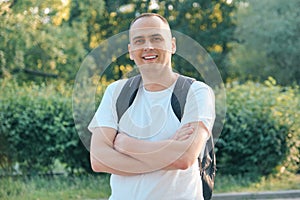 Outdoor portrait of mature smiling positive man. Confident male in white T-shirt with backpack with folded hands looking at the