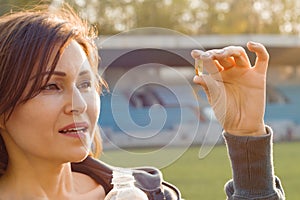 Outdoor portrait of mature woman taking vitamin E capsule pill of cod liver oil, at stadium. Sport, healthy lifestyle and