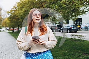 Outdoor portrait of modern young woman with mobile phone in the street. Happy redhead 30s girl texting on smartphone on