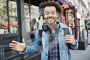 Outdoor portrait of positive african-american with afro hairstyle waving and smiling at camera while walking in street