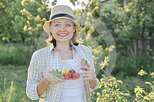 Outdoor portrait of positive mature woman in straw hat. Smiling female with plate of strawberries mint lemon, background green