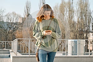 Outdoor portrait of pretty teen girl walking and texting on mobile phone, spring sunny day background