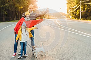 Outdoor portrait of pretty woman, her husband and daughter show ok sign, walk with dog on road, enjoy sunshine, have active lifest