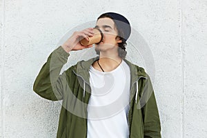 Outdoor portrait of relaxed youngster drinking hot drink from papercup outdoors, closing his eyes, feeling pleasure, being on