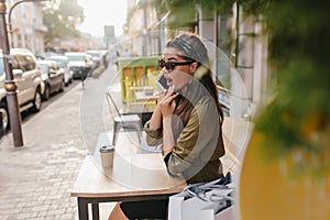 Outdoor portrait of shocked brunette girl in sunglasses talking on phone in autumn morning. Surprised shopaholic woman