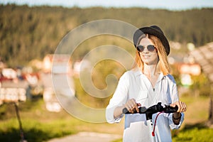 Outdoor portrait of stunning blonde girl in sunglasses on city on kick scooter. Enjoying sunny day with ecological transport