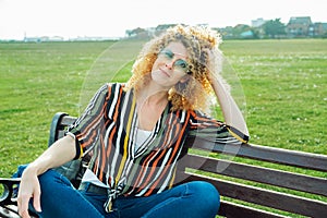 Outdoor portrait of stylish happy smiling curly hair woman wearing flower shaped sunglasses, striped multicolor shirt