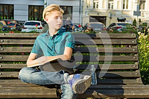 Outdoor portrait of teenage boy of 13, 14 years old sitting on bench in city park.