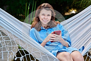 Outdoor portrait of teenage girl sitting in hammock with smartphone