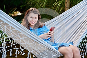 Outdoor portrait of teenage girl sitting in hammock with smartphone