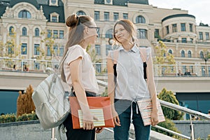 Outdoor portrait of two young beautiful girls students with backpacks and books talking and smiling, urban background