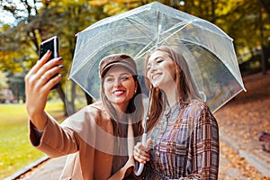 Outdoor portrait of two young women taking selfie using phone under umbrella during rain in autumn park