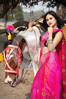 Outdoor portrait of very beautiful Indian girl wearing saree and holds the reins saddled horse and posing fashionable in blurred